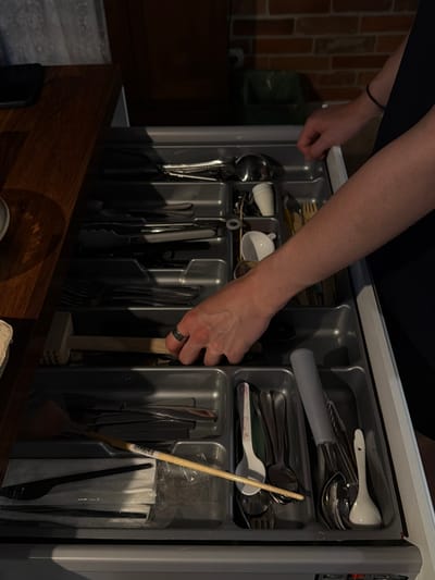 The image shows a person's hands reaching into an organized kitchen drawer filled with cutlery. The drawer has multiple compartments, each holding different types of utensils. We see forks, spoons, knives, and other kitchen tools neatly arranged. A light-colored ceramic spoon and a white ceramic cup are visible in one section. In another, a wooden handled utensil, possibly a small mallet or pestle, is propped up. The person's hand, adorned with a dark ring, is actively engaged in manipulating one of the utensils. The background suggests a kitchen setting, with a glimpse of a brick wall and a dark countertop. The lighting is somewhat dim, creating shadows that highlight the textures of the cutlery and the drawer's interior. The overall scene conveys a sense of order and domestic activity, likely the act of putting away or retrieving kitchenware. There is no visible text in the image.