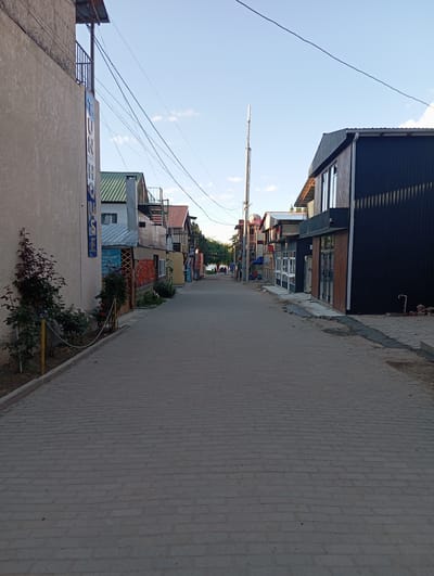 The image depicts a street scene in Bosteri, Kyrgyzstan. The street is paved with grey cobblestones and appears to be a pedestrian walkway or a narrow road lined with buildings. The buildings on either side are of varying architectural styles and colors, with some featuring multi-story structures and balconies. One building on the left has a sign that appears to say "TUNHOUSE". Power lines crisscross the sky above the street. In the distance, the scene suggests a transition towards a more open area, possibly a lake or a park, with trees and a few people visible. The sky is a clear blue with some scattered clouds, indicating daytime. The overall impression is that of a developing or resort area, with a mix of permanent structures and more modern establishments.