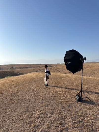 The image, taken outdoors in Sidi Badhaj, Morocco, shows a person standing in a dry, grassy field. To the left is a Profoto brand umbrella light modifier mounted on a stand. The person is wearing a white garment and appears to be holding an object. The background features rolling hills in the distance and a clear, light blue sky above. The terrain is mostly dry with sparse vegetation.