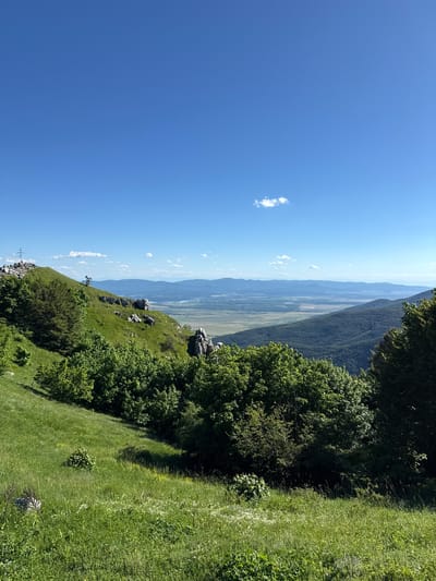 The image depicts a scenic mountain landscape with a clear blue sky. In the foreground, a grassy slope is covered in lush green grass and scattered wildflowers. Several trees are visible, with dense green foliage.
In the middle ground, a rocky outcrop juts out from the hillside, with some rocks also scattered on the grassy slopes. Further in the distance, a valley unfolds, dotted with greenery and a body of water, possibly a river or lake. The valley is framed by rolling hills and distant mountains, layered in shades of blue.
On the highest peak to the left, a cross is visible, suggesting a landmark or memorial. A few small, white clouds drift across the sky. The overall scene suggests a natural, elevated viewpoint, likely a hiking trail or a viewpoint overlooking the surrounding region.
While the image is described as being in the city of Shipka, Bulgaria, there are no specific man-made structures or urban elements directly visible in the frame that would definitively pinpoint the exact location within the city limits. The scene focuses on the natural beauty of the mountainous terrain. There are no people or animals immediately apparent in the image.
