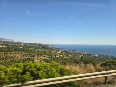 The image is a scenic view from a moving vehicle, likely a car, looking out over a coastal landscape under a clear blue sky. In the foreground, a blurred green shrubbery is visible, with a silver guardrail running horizontally across the lower portion of the frame. Beyond the guardrail, the landscape unfolds with rolling green hills dotted with trees and some scattered buildings. A winding road can be seen traversing the hills. In the middle ground, a town or village with red-tiled roofs is situated along the coastline, bordering a tranquil blue sea. The sea extends to the horizon, meeting the clear sky. The scene suggests a sunny day in a Mediterranean or similar coastal region, characterized by lush vegetation and a seaside settlement. The image is not from the Metropolitan Borough of Solihull, United Kingdom, as that location is inland and not coastal.