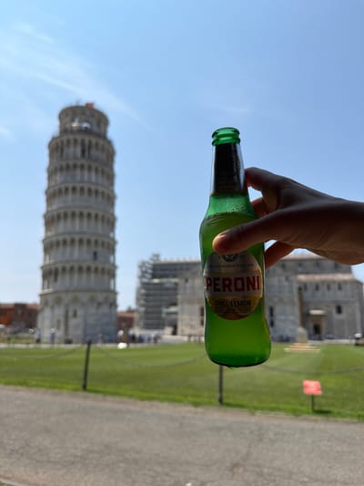 In the city of Pisa, Italy, a hand holds a green glass bottle of Peroni Chill Lemon beer in the foreground, with the iconic Leaning Tower of Pisa blurred in the background. The tower, a cylindrical bell tower, stands tall with its distinctive white marble construction and arched windows. Behind it, other buildings, possibly part of the Piazza dei Miracoli, are also visible, some with scaffolding. A grassy area with a chain-link fence separates the paved foreground from the buildings. The sky is a clear, bright blue, indicating a sunny day. A few indistinct figures can be seen in the distance, appearing to be tourists.