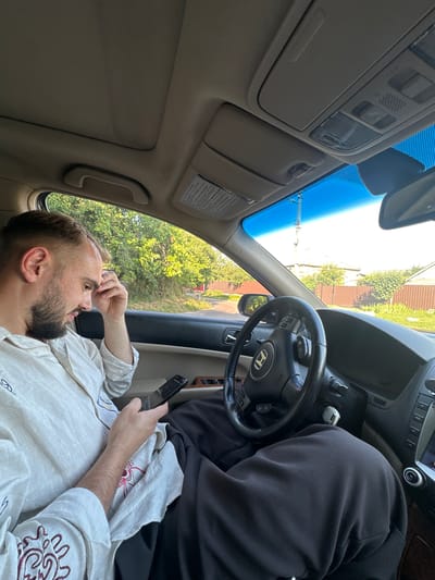 A man lies back in the passenger seat of a car, looking upwards and to the right with his hand on his forehead. He has a beard and is wearing a light-colored, loosely woven shirt. He is holding a black smartphone in his other hand, which is resting on his lap. A person wearing a black hooded garment is seated in the driver's seat, facing forward, with their head obscured by the hood.
The interior of the car is visible, including the steering wheel and dashboard. The sunroof is open, revealing a bright, clear sky and trees outside. The sun appears to be high in the sky, suggesting it is daytime. The scene appears to be set in a residential area, with a fence and houses visible through the open sunroof and side window. The general atmosphere is relaxed and casual. No visible text is present in the image.