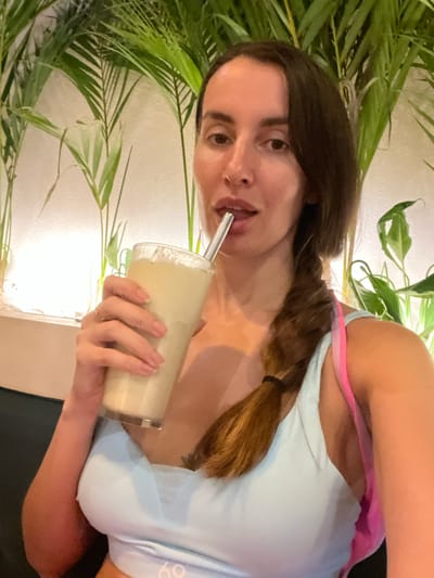 A woman with dark brown hair in a braid and wearing a light blue sports top with pink straps is drinking from a tall glass with a metal straw. She is indoors, surrounded by green tropical plants. Based on the prompt, the location is Pererenan, Indonesia.