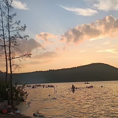 The image depicts a lake scene at sunset in Zerendi, Kazakhstan. In the foreground, a sandy shore is visible with several pairs of flip-flops, a red ball, and some discarded plastic bags. Two bare trees stand on the left side of the frame. The lake itself is populated with numerous people swimming and floating on inflatable rings. Some individuals are standing in the water, while others are partially submerged. The water reflects the warm hues of the sunset, casting an orange glow. In the background, a dark, tree-covered hill forms the horizon. The sky above is a gradient of pale blue to warm orange and pink, with scattered clouds illuminated by the setting sun. The overall atmosphere is one of leisure and relaxation on a summer evening.