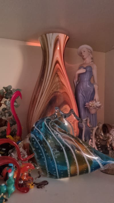 A decorative shelf displays various glass figurines and a statue. In the center, a tall, swirled vase with warm tones of brown and orange stands prominently. To its right, a porcelain figurine of a woman in a purple dress and white hat holds a basket of flowers. In the foreground, a blue and green swirled glass object, possibly a bowl or sculpture, is positioned at an angle. To the left of the vase, a bright pink, curved glass piece, resembling a flamingo neck, emerges from a cluster of green glass leaves. Several smaller, colorful glass figurines, including what appear to be small animals or mythical creatures, are scattered in the foreground and around the base of the larger glass object. The background is a plain, light-colored wall, and the scene is lit from above, casting a warm glow on the objects. The overall impression is one of an eclectic collection of decorative items. This image was taken in Aleksin, Russia.