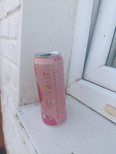 A pink aluminum can of energy drink "DRIVE SAMOOBRAZhenIYA" with text in Russian is placed on a white windowsill. The can is tilted slightly to the right. The background shows a white brick wall and a white window frame. The image appears to be taken outdoors. The location is Krutinka, Russia.