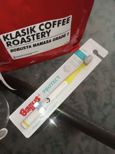 A red bag of "Klasik Coffee Roastery Robusta Mamasa Grade 1" is positioned beside a packaged "Bagus Protect" toothbrush. The toothbrush, yellow and white, is labeled "soft" and "care + natural" in English and Chinese. It's in a white and clear plastic package. The background is a dark, reflective surface.
