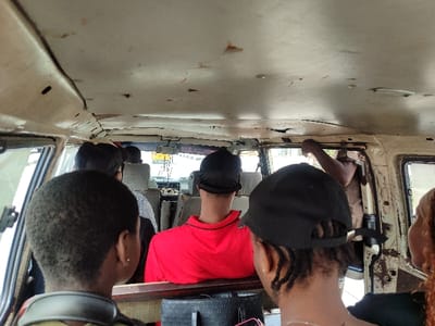 The image depicts the interior of a passenger van or minibus in Igando Oloja, Nigeria, viewed from the rear passenger area looking forward. The vehicle's ceiling is light-colored, showing significant wear, discoloration, and multiple small holes or indentations.

Approximately six individuals are visible inside the vehicle. In the foreground, the back of a person's head with short, dark, closely cropped hair is seen on the left, wearing what appears to be a light-colored strap around their neck. To the right of this person, another individual's head is visible, wearing a black baseball cap facing backward, revealing dark dreadlocks at the nape of the neck. Further ahead, a person wearing a red shirt is seated, facing forward, with their back to the viewer. Beyond them, other passengers are partially obscured, with one wearing a striped shirt visible.

On the right side of the vehicle, near a doorway or large window opening, a person's dark-skinned arm is raised, bent at the elbow, and appears to be holding onto a part of the vehicle's frame. The interior structure of the vehicle exhibits signs of wear and age, with visible metal frames and unadorned surfaces. Through the front windshield, a blurry view of an exterior environment, possibly a street, is visible, including what appears to be a yellow vehicle in the distance. The scene suggests a public transport setting.