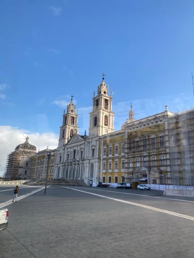 The image captures a wide view of the Mafra National Palace in Mafra, Portugal, under a clear blue sky with sparse white clouds. The grand, Baroque-Neoclassical structure occupies the midground and background, featuring a prominent central basilica flanked by two tall, light-colored bell towers with decorative spires and visible bells. A large, paved plaza of dark grey pavers stretches across the foreground and midground.
The palace complex extends horizontally, with a yellowish facade on the right side and a lighter facade on the left. Significant portions of the building are undergoing renovation; the right wing and a dome structure on the far left are covered in extensive scaffolding. A wide set of stairs leads up to the main entrance of the central basilica.
In the plaza, a single individual is walking towards the left in the lower-left quadrant, wearing a brown jacket and light-colored pants. A white pickup truck is partially visible in the bottom-left corner. Near the base of the renovated right wing, several vehicles, including multiple trucks and a white car, are parked behind white barriers. A tall, dark pole, possibly a street light, stands in the midground of the plaza. Subtle reflections on the image surface suggest it may have been captured through a window.