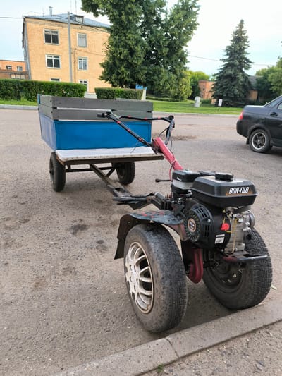 This media file captures a stationary two-wheeled tractor with a trailer attached, parked on an asphalt road in what appears to be a semi-rural or suburban setting. The tractor is red and black, featuring a prominent "DON-FILO" engine. The trailer is blue with wooden sides and a white upholstered seat.
The scene is set outdoors, likely during the daytime, with overcast skies suggesting mild weather. In the background, a two-story brick building with multiple windows is visible, along with mature trees and a dark-colored sedan parked on the right side of the frame. The ground is paved with asphalt and there is a concrete curb in the foreground.
No people are present in the image, and the tractor and trailer appear to be idle. The overall impression is one of functional utility, suggesting the equipment is either parked after use or awaiting operation. The location context provided is Uzunovo, Russia. The text visible on the engine is "DON-FILO" and "212 cc".