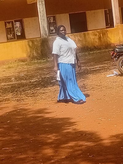 A woman stands in a schoolyard, dressed in a white shirt and long blue skirt. She holds a white paper. Nearby, a building with windows and mounted items is visible. The ground is a dry, reddish-brown color with sparse vegetation. Dark shadows are cast on the ground, possibly from trees or other structures. A motorcycle is parked near the top of the frame. The overall impression is of a rural or less developed setting.