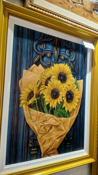 The image is a close-up, angled shot of a framed oil painting featuring a bouquet of sunflowers. The sunflowers, predominantly yellow with dark centers, are wrapped in brown paper. The background of the painting is a dark blue, textured surface with ornate gold and black decorative elements, resembling a carved wooden door or panel. The painting itself is enclosed in a thick, ornate gold frame, with a white matte border. The overall presentation suggests an art gallery or a home displaying artwork, but there is no specific information to pinpoint its location in the city of Nyamat, Indonesia.