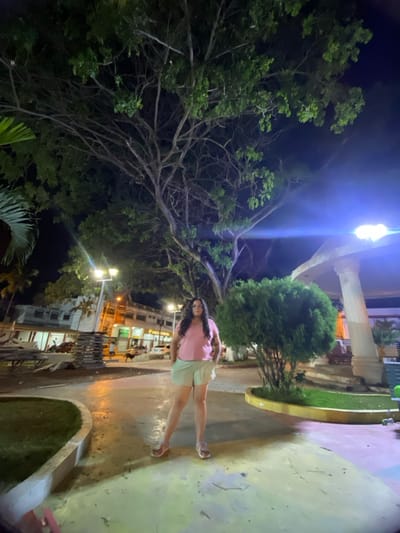 The image depicts an outdoor night scene in a public park or plaza in General Villamil Playas, Ecuador. An adult female stands centrally in the foreground, facing the camera with hands in pockets. She has long, dark, wavy hair, wears glasses, a pink t-shirt, light-colored shorts, and sandals. Behind her, a very large tree with extensive branching and green foliage dominates the upper frame, illuminated by artificial light. To the woman's right, a dense, green shrub is present, and a white gazebo-like structure with a prominent column is partially visible further right.
The ground is paved, showing varied colors, and a curved, low-walled grass area is to the left. Multiple streetlights on poles illuminate the scene, casting bright light and causing a lens flare on the right. In the background, across a street, a row of multi-story commercial buildings is visible, some with illuminated signs, including text resembling "Puerto Pesquero." Parked vehicles and indistinct figures of people are present on the street. To the left, near the street, a dark-colored dog is visible, along with what appears to be stacked or disassembled picnic tables or benches. Palm fronds are visible on the far left.