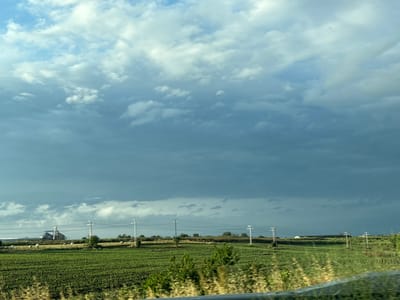 The image depicts a rural landscape under a dramatic, cloudy sky, likely taken near Chirpan, Bulgaria. The foreground features a vibrant green field with rows of crops, bordered by dry, tall grass. In the mid-ground, a line of white hay bales is scattered across a field of golden stubble. Power lines supported by tall, white pylons stretch across the scene, with some smaller, dark poles also visible. In the distance, a cluster of industrial buildings, possibly grain silos, stands against the horizon. The sky is a mix of bright, white clouds and darker, ominous storm clouds, suggesting changing weather conditions. The overall impression is one of an agricultural area experiencing dynamic weather.