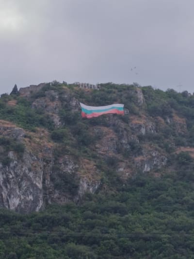 The image shows a large Bulgarian flag draped across a rocky hillside. The flag is positioned prominently on the slope, with its white, green, and red stripes clearly visible. Above the flag, large white letters spelling out "КРИЧИМ" (Krichim) are built into the side of the hill. The hillside is covered in sparse vegetation, with rocks and earth exposed in places. The foreground of the image is filled with dense green trees, suggesting a forested area below the hill. The sky is overcast with a grey, diffused light, indicating it is likely daytime but not a sunny day. The overall impression is one of national pride and a landmark visible from a distance. The time of day appears to be overcast daylight, and the location is identifiable by the prominent name of the town.