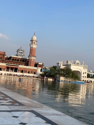 The image depicts the Golden Temple complex in Amritsar, India. The scene is dominated by the reflective waters of the sarovar (pool) in the foreground, with the iconic architecture of the temple and its surrounding buildings visible across the water.
In the background, a tall, red and white striped minaret pierces the clear blue sky, a prominent feature of the complex. Adjacent to it, a building with arched windows and a white dome is visible. To the right, a grand white structure with multiple domes and arches stands out, reflecting in the water. A lush green tree partially obscures this white building.
A large number of people are gathered along the edge of the sarovar, some standing, others appearing to be bathing or walking. Their presence suggests a pilgrimage or a visit to this sacred site. The water itself is calm, with ripples creating a distorted reflection of the surrounding architecture. The foreground features a patterned marble walkway, likely part of the perimeter of the sarovar.
The overall impression is one of serenity and spiritual significance, typical of the Golden Temple. The clear sky and the calm water contribute to a peaceful atmosphere.