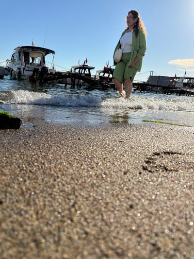 The image captures a woman walking through shallow water near a beach in Üsküdar, Turkey. She is wearing a light green suit and a white top, with a cream-colored bag slung over her shoulder. Her feet are splashing in the water, creating ripples. In the background, several boats are docked near a wooden pier. The sky is clear and blue, with the sun setting, casting a warm glow on the scene. The foreground is dominated by a close-up view of wet sand, reflecting the sunlight.