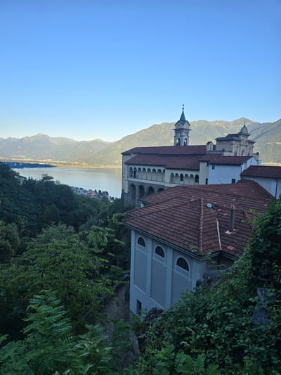 The image depicts a scenic view of the Madonna del Sasso sanctuary in Orselina, Switzerland. The sanctuary, with its distinctive tiled roofs and bell tower, is situated on a hillside overlooking a serene body of water, likely Lake Maggiore. In the background, a range of mountains stretches across the horizon under a clear blue sky. The foreground is dominated by lush green trees and vegetation, with the stone facade of a building also visible. The overall impression is one of tranquility and natural beauty, with the sanctuary serving as a focal point within the landscape.