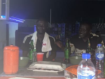 Two men are seated at a table outdoors at night. The man on the left has a white towel draped around his neck and is looking forward. The man on the right is smiling and looking to his left. On the table in front of them are several bottles of beer, a bottle of water, a red cup, a green bowl, and an orange container. In the background, there are stacks of what appear to be sound equipment and a fence with barbed wire. The scene is lit by artificial lights, including blue lights on a structure to the left. The setting is consistent with an outdoor gathering in Bwari, Nigeria, likely in a residential or entertainment area.