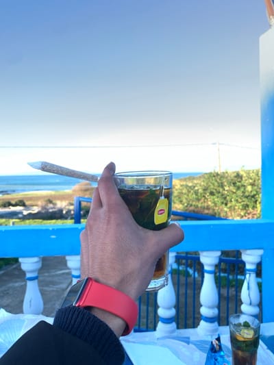 A hand holds a glass of tea with a Lipton teabag, alongside a rolled cigarette. The person wears a red Apple Watch band. In the background, blue painted posts and white spindles are visible, with glimpses of greenery, sky, and a body of water.