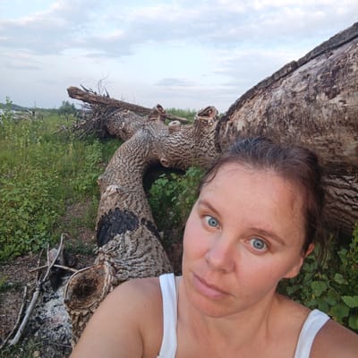 A woman is captured in a selfie in a rural area of Старый Буртюк, Russia. She is in the foreground, with her face and upper chest visible. Behind her, a large, fallen tree trunk dominates the scene, its bark textured and weathered. The trunk is partially covered in green foliage. To the left of the trunk, there is a patch of dry, burnt wood and ash, suggesting a past campfire. The background consists of a field of green grass and some scattered branches, under a cloudy sky. The overall impression is one of a natural, somewhat wild environment.