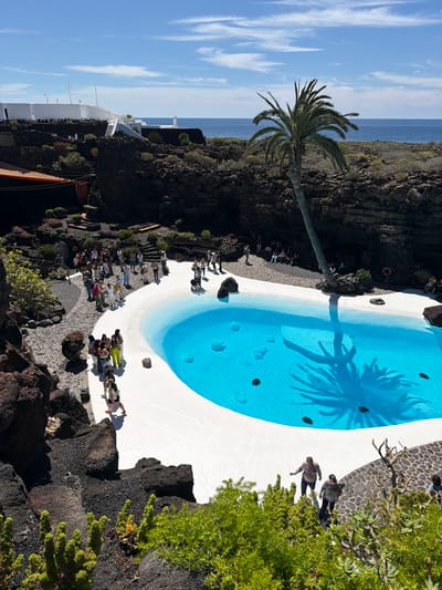 The image captures an elevated view of a unique pool complex at the Casa Museo César Manrique in Haría, Spain. A turquoise blue swimming pool dominates the center, with a prominent shadow of a palm tree stretching across its surface. The pool's perimeter is painted white, blending into the surrounding rock landscape.

Several individuals are present. A group stands near a paved area next to the pool, while two people are seen walking near the edge.

The environment is characterized by volcanic rock formations, indicative of Lanzarote's geological landscape. The pool is located near the coast, with a glimpse of the ocean visible in the background under a blue sky.