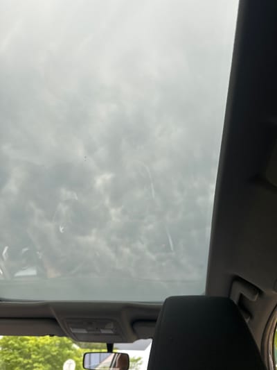 The image is taken from inside a vehicle, looking upwards towards the sky through a sunroof or windshield. The sky appears overcast with diffuse sunlight, suggesting it is daytime. The clouds are a prominent feature, creating a mottled pattern of light and dark grey. Reflections are visible on the glass, showing glimpses of the vehicle's interior, including parts of the roof lining, a sun visor, and potentially other passengers or objects within the car. A side-view mirror is also visible, reflecting a green, leafy environment, likely trees or foliage outside the vehicle. The overall impression is one of a travel scene, possibly on a cloudy day. There is no visible text in the image.