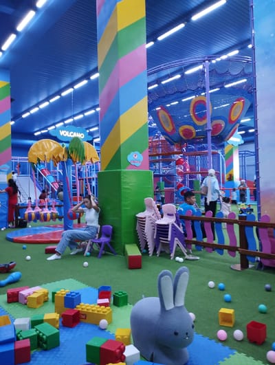 This is a candid, eye-level, indoor photograph of a brightly colored children's play area, possibly in Talas City, Kyrgyzstan. The environment is a large, open space with a high blue ceiling illuminated by rows of fluorescent lights. A prominent, large, cylindrical pillar painted with diagonal stripes of yellow, green, pink, and blue stands in the foreground to the left of center. To the right of this pillar is a wall covered in a green padded material.
In the mid-ground, a young woman in a white t-shirt and blue jeans sits on a small purple chair, with her hands on her head, appearing somewhat distressed or tired. In front of her are large, colorful building blocks in red, yellow, green, and blue, scattered across a green mat. A large, gray, stuffed rabbit toy is positioned in the lower right quadrant of the image.
The background reveals a multi-level play structure with slides, nets, and climbing areas in various colors, including red, blue, and yellow. Several other children and adults are visible, interacting within the play area. Hanging from the ceiling above the play structure is a sign that reads "VOLCANO" in white lettering on a blue cloud-shaped background. The floor is covered in artificial green turf and padded mats. The overall impression is a lively and energetic indoor playground.