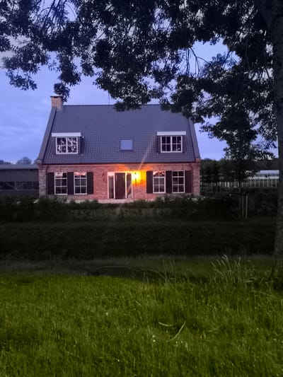 The image depicts a detached brick house with a dark gray tiled roof, seen at dusk or dawn. The house has multiple windows, some with decorative white grilles on the upper floor and black shutters on the lower floor. One window on the left of the upper floor has a dormer. A prominent orange light emanates from the front door area, illuminating the entrance and the adjacent window. A chimney made of red bricks is visible on the roof. The house is surrounded by dense, dark green foliage and trees. In the foreground, a strip of green grass is visible, with some taller blades swaying. The background shows more greenery and what appears to be a greenhouse structure to the right of the house, and a larger building or shed-like structure to the left, partially obscured by trees. The sky is a deep blue, indicative of twilight. The image is taken from a slightly elevated position, possibly across a field or body of water. The overall impression is a quiet, rural or semi-rural setting. The house is located in Almere, Netherlands.