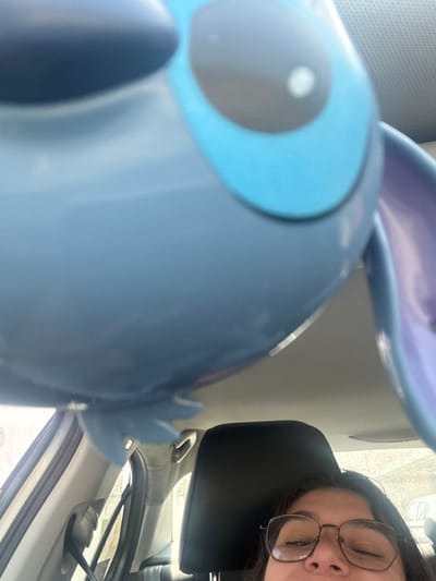 A close-up image taken from inside a vehicle depicts a large, light blue object, identifiable as the character Stitch from Disney's Lilo & Stitch, occupying the upper portion of the frame. The Stitch object is out of focus, showing its left eye with a black pupil, bright blue iris, and a white specular highlight, along with a segment of its purple ear on the right. In the lower right quadrant, a human subject, possibly a female with dark hair, is partially visible. She wears glasses with thin, metallic frames, and her eyes are partially closed or squinting. The vehicle's interior includes a black headrest behind the person and light-colored plastic trim components along the ceiling and left pillar. Through the vehicle's windows, a blurry outdoor environment under overcast lighting is visible, with indistinct shapes of structures on the left and a light-colored vehicle partially seen on the far right. The specified location of Casa Blanca, Mexico, cannot be confirmed through visual evidence within the image.