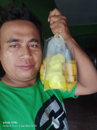 A smiling man in a green shirt holds up three plastic bags filled with yellow food and liquid. The bags appear to contain tofu and broth. The man has dark hair and fair skin. The photo was taken with a Vivo Y19s camera on December 2, 2025, at 7:45 AM, according to the watermark. The background is blurred, but a glimpse of a tile floor is visible.