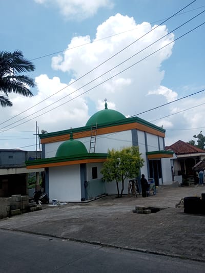 The image shows a two-story building with white walls, green domes, and an orange stripe. It's likely a mosque, given the domes and architectural style. A ladder is leaning against the building. A person stands next to a food cart near the building's entrance. A tree is also located near the entrance. A motorcycle is parked nearby another building. The foreground consists of a concrete area with some uneven surfaces and a portion of a street. Power lines are visible against the blue sky with white clouds. There is no evidence that the image is located in Xayabury District, Laos.