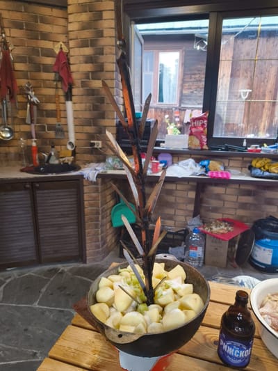 The image shows an outdoor kitchen or patio area, possibly in Востец Снт, Russia. In the foreground, a large metal pot filled with cubed potatoes and some green ingredients is placed on a wooden table. A metal spit with multiple prongs extends vertically from the center of the pot, suggesting food preparation. To the right of the pot, a dark brown glass bottle with a label reading "РИЖСКОЕ" is present. Further to the right, a white bowl contains raw meat. The background reveals a brick wall, kitchen cabinetry, a window with a view of a wooden building, and shelves with various items like a bag of chips and plastic containers. The overall scene suggests a setting for outdoor cooking or a barbecue.