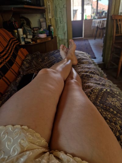 In Raduvene, Bulgaria, an individual's legs are prominently displayed, resting on a patterned blanket and an orange and black plaid blanket, atop what appears to be a couch or bed. The person is lying down, with their feet visible at the top of the frame. Their legs are crossed, and they are wearing light-colored, patterned underwear or loungewear at the bottom of the image.
The background suggests an indoor setting, possibly a living area. To the left, a wooden shelf holds various items, including bottles and boxes. The wall behind the shelf has a textured, light-colored finish. To the right and in the background, an open doorway leads to an outdoor or semi-outdoor space, revealing tables, chairs, and a glimpse of a building and sky. The overall scene has a casual, domestic atmosphere.