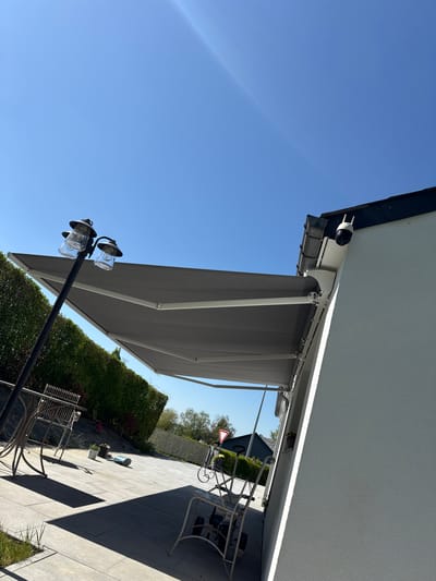 The image shows an outdoor patio area in Marche-en-Famenne, Belgium. A retractable awning extends from the side of a light-colored building, providing shade. Patio furniture, including chairs and a small table, are visible on the paved patio. A light post with two glass-enclosed fixtures stands near the edge of the patio. A neatly trimmed hedge forms a boundary to the patio area. In the background, a residential street with a traffic sign can be seen. The sky is clear and blue. A security camera is mounted on the building near the awning.