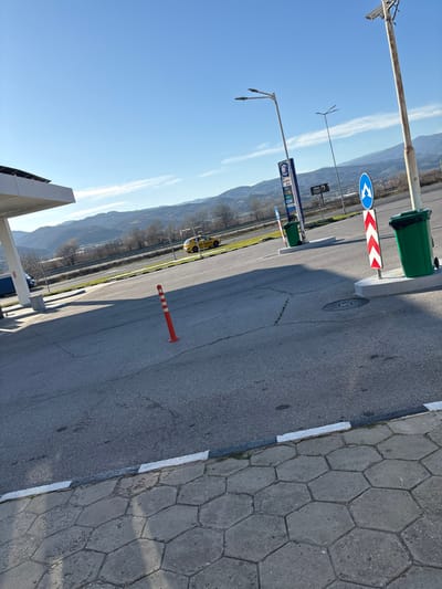 The image depicts a roadside service area on a clear, sunny day, identified as being in Strumyani, Bulgaria. The foreground features a sidewalk paved with grey hexagonal blocks, separated by a curb with a white stripe from an asphalt surface exhibiting minor cracking. An orange traffic cone with white reflective stripes stands upright on the asphalt in the mid-left.
In the midground, the edge of a white service station canopy is visible on the far left, partially obscuring a dark blue truck. Beyond the immediate asphalt area, a multi-lane road stretches horizontally, upon which a yellow hatchback car is in transit, moving from left to right. Further right, two streetlights of differing designs are present; one features a double-arm fixture and solar panels, while the other is a simpler, taller pole. Between these, a tall vertical sign, possibly a gas station price board, displays "Кафе" (Coffee) and other text. Two green waste bins, each on a concrete base, are positioned in the right midground. Adjacent to the rightmost bin, a blue circular road sign with a white left-pointing curved arrow is mounted above a red and white chevron sign. A round metal manhole cover is visible on the asphalt near the right green bin.
The background consists of a low strip of bare trees and foliage, beyond which a range of undulating hills and mountains extends under a clear blue sky with minimal cloud cover. No individuals are explicitly visible, and no direct interactions are depicted other than the implicit presence of a driver in the moving yellow vehicle.
