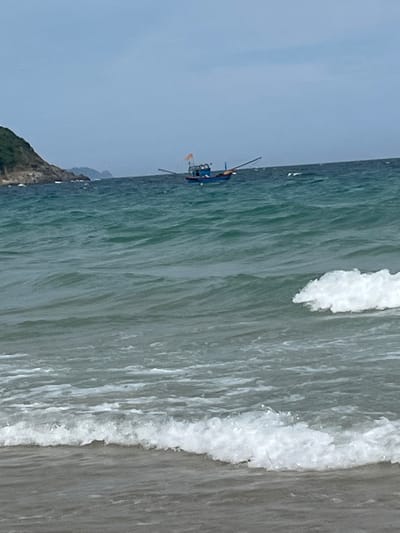The image shows a small blue and orange fishing boat with a single pole in the water. The boat is in the ocean, with waves crashing on the shore to the right of the frame. A rocky, tree-covered coastline is visible in the background on the left. The sky is a pale blue, indicating it is likely daytime. The scene appears to be a calm day at sea, possibly in Cam Lâm, Vietnam. There is no visible text in the image.
