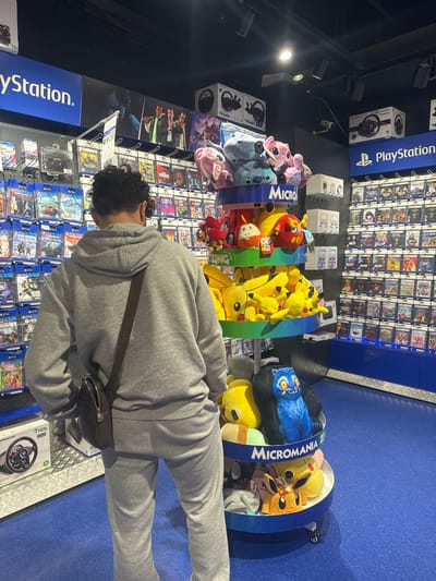 A person wearing a grey hoodie and sweatpants stands in a store, facing away from the camera. They have a dark brown messenger bag slung across their body. The person is in an aisle of a retail store, likely a video game store, with shelves of video games visible on the right side of the image. In front of the person, there is a multi-tiered display stand filled with plush toys, predominantly featuring characters from popular gaming franchises like Pokemon. The store appears to be a Micromania store, as indicated by branding on the display. The background shows more rows of video games and store signage, including a large "PlayStation" logo. The floor is a vibrant blue. The location is identified as Leers, France.