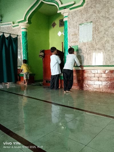 The image depicts three young boys standing inside a mosque, likely in the town of Anjatan, Indonesia. The mosque features tiled walls, a green tiled ceiling, and a single, large green curtain. The boys are all wearing casual attire: one in a yellow shirt and shorts, another in a white shirt and black pants, and the third also in a white shirt and black pants. Two boys stand on the right, facing a wall, and looking up. The boy on the left is looking forward, with a wooden podium to his right. The scene is lit by natural light coming in from a window, possibly an arched doorway. There are reflections of the boys on the tiled floor. The atmosphere is peaceful, and the boys appear to be engaged in quiet contemplation.