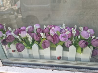 The image displays a window box filled with artificial purple and pink flowers, accented by small white floral sprigs. The flowers are arranged behind a miniature white picket fence. Two ladybugs are visible on the fence; one is on the left side, and the other is on the right. The window box appears to be placed on a windowsill. Reflections in the glass suggest an outdoor urban environment, possibly a street scene with buildings and vehicles in the background, though these are blurred. The overall impression is a decorative arrangement, likely intended to beautify a window or balcony. No specific location within Antony, France can be determined from the image alone.