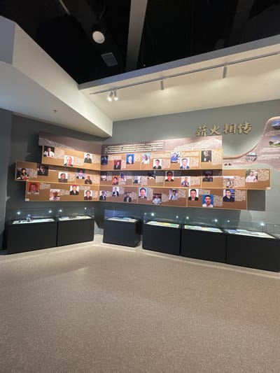 An interior space features a wall display of framed portraits and text, organized vertically. To the right, black rectangular display cases are arranged sequentially. The floor is a uniform light gray. The left wall has a white section with visible lighting fixtures and a darker, glossy area.