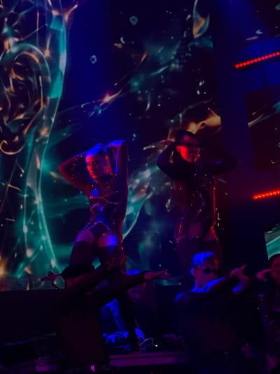 The image depicts a stage performance in Tsilkani, Georgia, characterized by vibrant lighting and a futuristic backdrop. Two female performers, dressed in shimmering, dark, form-fitting outfits, are the central focus. The performer on the left has her hands raised above her head, with her head tilted down. The performer on the right is also raising her hands, with her head tilted and mouth open, possibly singing or speaking into a microphone. Both performers have dark hair styled in ponytails. In the foreground, several other individuals are visible, also appearing to be part of the performance or crew, with one person in the center wearing sunglasses. The background features a large, abstract, glowing digital display with swirling green, blue, and orange hues, creating an immersive and dynamic atmosphere. Red LED lights are mounted above and to the right of the stage. The overall scene suggests a contemporary music or dance event.