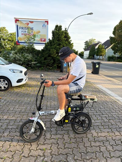 This media file shows a young person on an electric bicycle on a paved sidewalk. The person is wearing a black bucket hat, a white t-shirt with grey striped shorts, and white athletic shoes. They are holding the handlebars of the bicycle and looking down at a phone in their hand. The electric bicycle is white with black accents and has "MIDNIGHT HITWAY" written on the frame.

The setting appears to be a street in Nienburg/Weser, Germany. Behind the person and bicycle, there is a road with two trash cans and some trees and houses in the background. To the left of the frame, there is a large billboard advertising Aldi products, specifically cheese, with prices shown. The weather appears to be sunny, casting shadows on the ground. The time of day seems to be late afternoon or early evening, judging by the angle of the sun and the lighting. The overall mood seems casual and everyday.