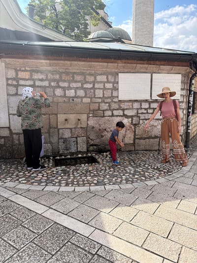In Sarajevo, Bosnia and Herzegovina, outside a historic building constructed of rough-hewn stone blocks, three individuals are present near an outdoor water fountain. A person wearing a patterned headscarf, green floral-print jacket, and black pants stands on the left, drinking from a clear bottle. To the right, a young boy in a grey t-shirt and red pants is bending down, appearing to interact with the water. A woman in a pink t-shirt, a wide-brimmed hat, and patterned brown pants stands behind the boy, pointing towards the ground. The ground is paved with cobblestones. Behind the individuals, a stone wall features two white plaques with inscriptions, and a historical minaret rises in the background under a partly cloudy sky.