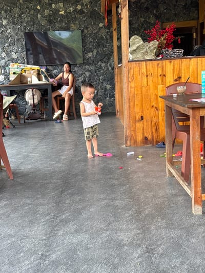The image captures a casual indoor or semi-outdoor scene, likely a cafe or restaurant in Du Già Commune, Vietnam. The setting features a distinctive mix of natural materials: dark grey, textured stone walls on the right and in the background, contrasting with warm-toned wooden panels and furniture on the left. The floor is dark grey, possibly slate or concrete, with visible texture.

In the bottom right of the frame, a woman with dark hair is seated on a low stool at a black table. She wears a brown tank top and white shorts, and appears relaxed, looking towards the left side of the frame while holding a small object in her hand. Beside her, a large, dark abstract painting hangs on the stone wall. The table is cluttered with various items, including a skateboard, a white baby chair, a fan, and other boxes or containers.

Towards the center of the image, a young child, possibly a boy, stands barefoot on the dark grey floor. He is wearing a white tank top and patterned shorts, and is holding a small orange box or container. The text "FOOD ME" is visible on his shorts. The child appears to be playing or casually observing his surroundings. Scattered on the floor around him are small, colorful toys, including a pink spade.

In the upper left, a wooden table and chairs are visible. On a wooden shelf or cabinet behind the table, decorative elements include a large grey rock and a cluster of artificial red berries. A small white sticker on the berries reads "SALE". The overall atmosphere is calm and domestic, suggesting a leisurely daytime moment.