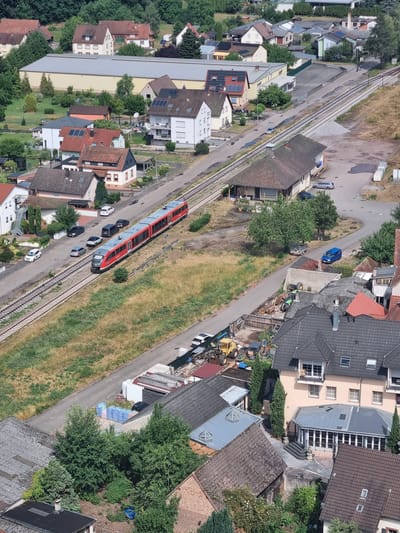 This aerial view depicts a train, likely a regional passenger train, traveling on a railway line that runs through a suburban or semi-rural area. The train is red and silver and appears to be moving from left to right in the frame. Several cars are parked along a road that runs parallel to the tracks. The scene is populated with residential buildings, some with red tile roofs and others with modern, flat roofs. There are also some commercial or industrial buildings with large, light-colored roofs. Green trees and vegetation are interspersed throughout the landscape, with dry, grassy areas suggesting a warm, possibly dry season. The time of day appears to be daytime, with clear lighting casting shadows. The overall impression is one of a quiet, functional community with transportation infrastructure integrated into the residential areas. No visible text is present in the image. The context suggests this location is Dahn, Germany.