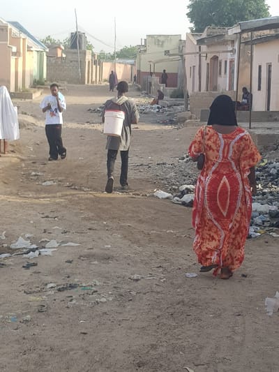 The image shows a street scene in what appears to be the Dutsinma B area of Nigeria. The road is unpaved, and littered with trash. Buildings line both sides of the street, showing signs of wear and limited infrastructure. Several people are visible: a woman in a patterned orange dress and a dark head covering walks toward the center of the image, apparently near a pile of trash. A man walks down the street carrying a white bucket. A young man walks down the street in a white t-shirt with dark pants. Other people are seen further down the street.