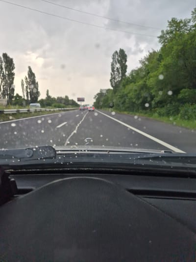The image shows a view from inside a vehicle looking out through a rain-streaked windshield onto a highway. Tall, slender trees line the right side of the road, and some trees are also visible on the left, interspersed with what appears to be a guardrail and a white vehicle. The sky is overcast and gray, with visible raindrops on the windshield. In the distance, several cars are on the highway, and a billboard is visible. The foreground of the image shows part of the vehicle's dashboard and windshield wiper. The scene appears to be a highway with moderate traffic, possibly in a rural or semi-rural area, with a damp, rainy atmosphere. The location is specified as the city of Hrazdan, Armenia.