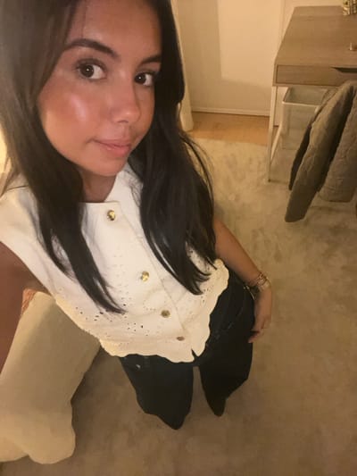 An indoor, vertical, full-body image portrays an adult female from a high-angle perspective, appearing as a self-portrait. The subject possesses long dark hair and wears a white, sleeveless top featuring eyelet embroidery and four visible gold-toned buttons, paired with dark trousers. Multiple bracelets adorn her left wrist, and a ring is visible on her left ring finger. She looks directly at the camera with a neutral to slight smile. The environment consists of a room with a light-colored carpet covering most of the floor, transitioning to light wooden flooring in the upper right. A light-colored wooden desk with a single drawer and a white metal frame is positioned in the background to the right. A square white bin is located beneath the desk. A quilted, olive-toned garment hangs draped over an object behind the desk. A light-colored upholstered item or cushion is partially visible on the left. Illumination is soft and warm, indicative of artificial indoor lighting. The precise location within Lordelo do Ouro e Massarelos, Portugal, is not discernible from the visual content.